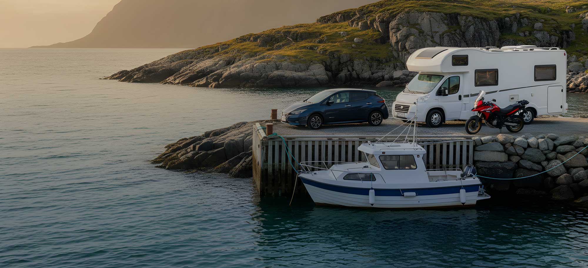 En bobil, en bil og en motorsykkel står parkert på en brygge ved sjøen, med en liten båt fortøyd i vannet og fjellandskap i bakgrunnen i kveldslys.