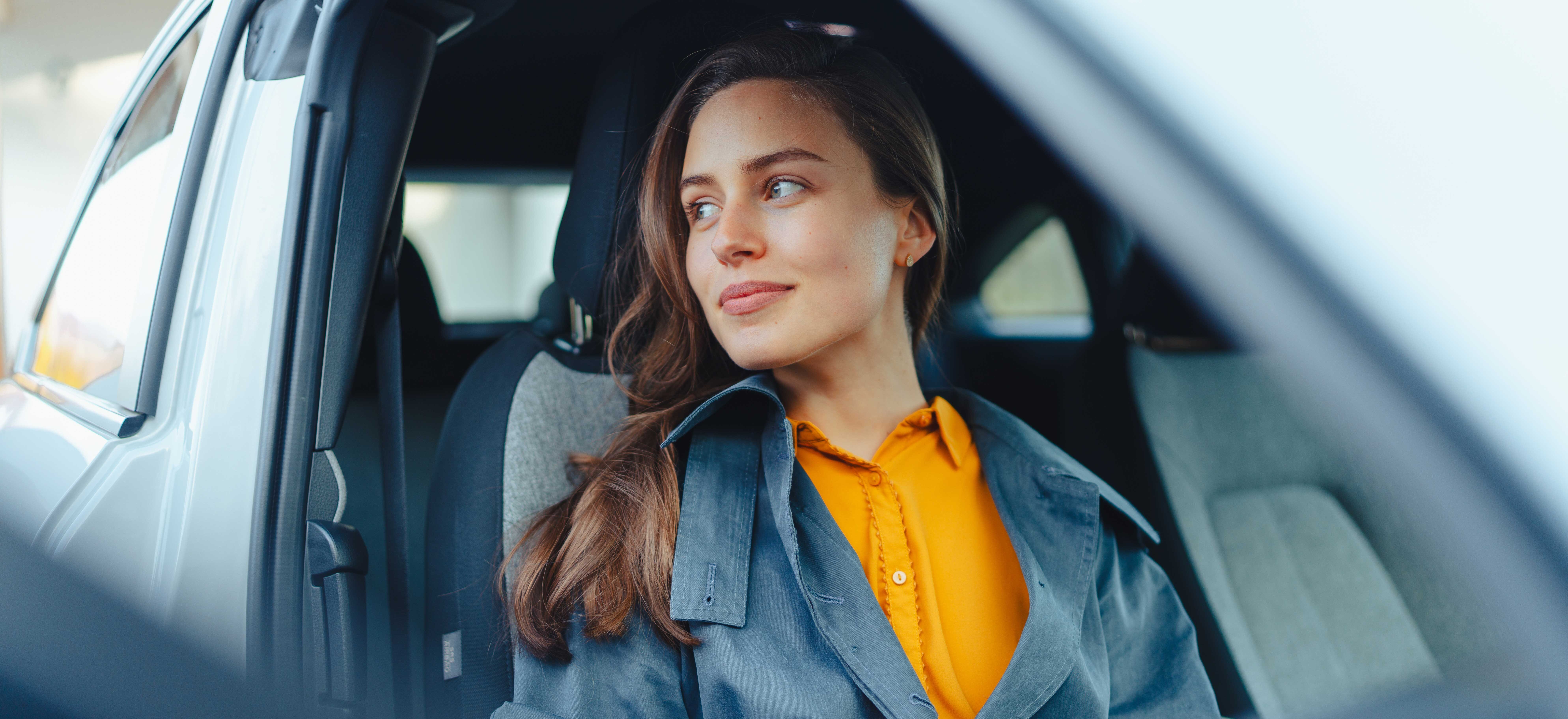 En kvinne med brun jakke og oransje skjorte sitter i bilen og ser ut av vinduet med et rolig uttrykk.