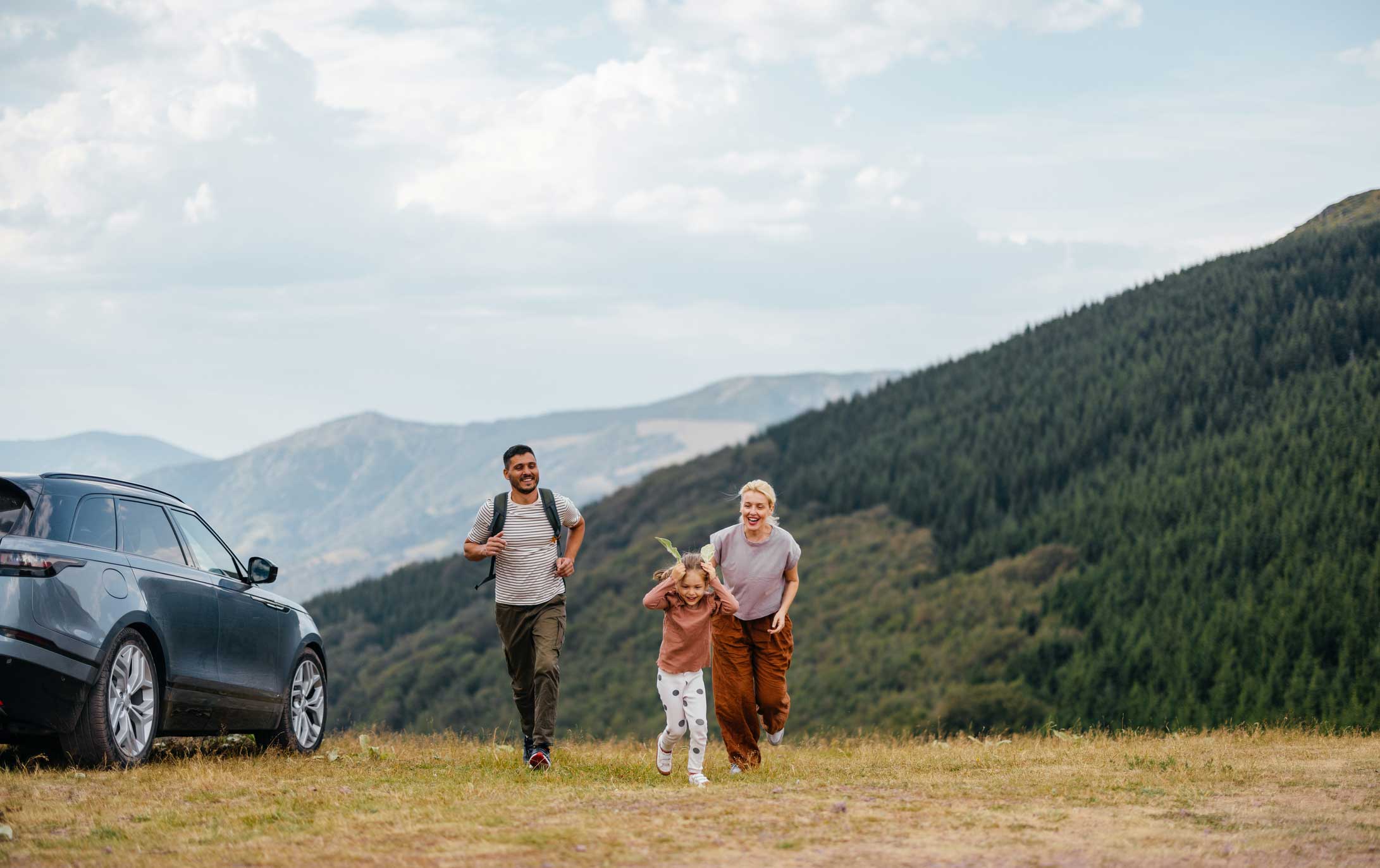 En familie med to voksne og et barn løper glade sammen i fjellandskapet ved siden av en parkert bil, med grønne åser og blå himmel i bakgrunnen.